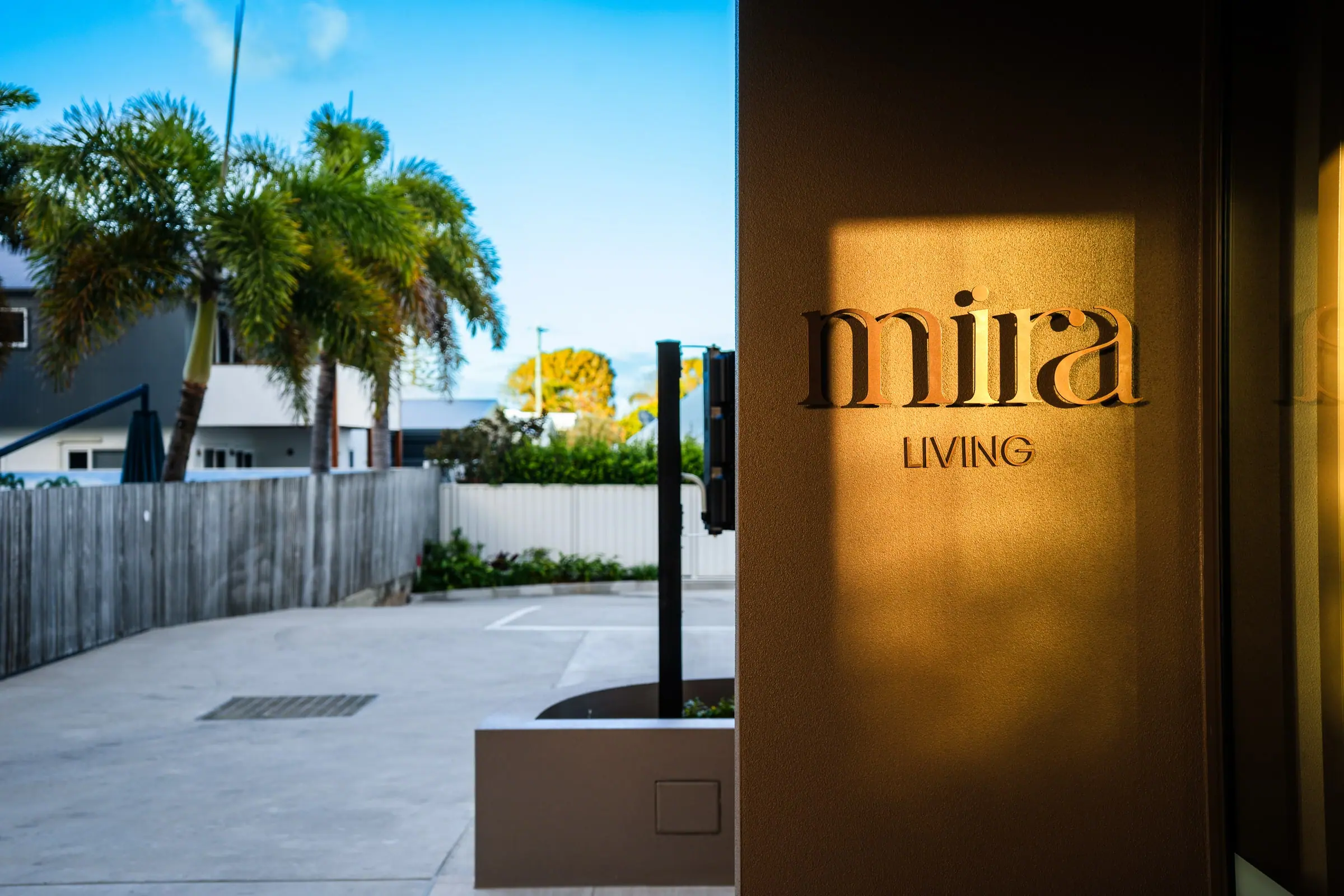 The Mira Living entrance at golden hour, palm shadows falling across the signage